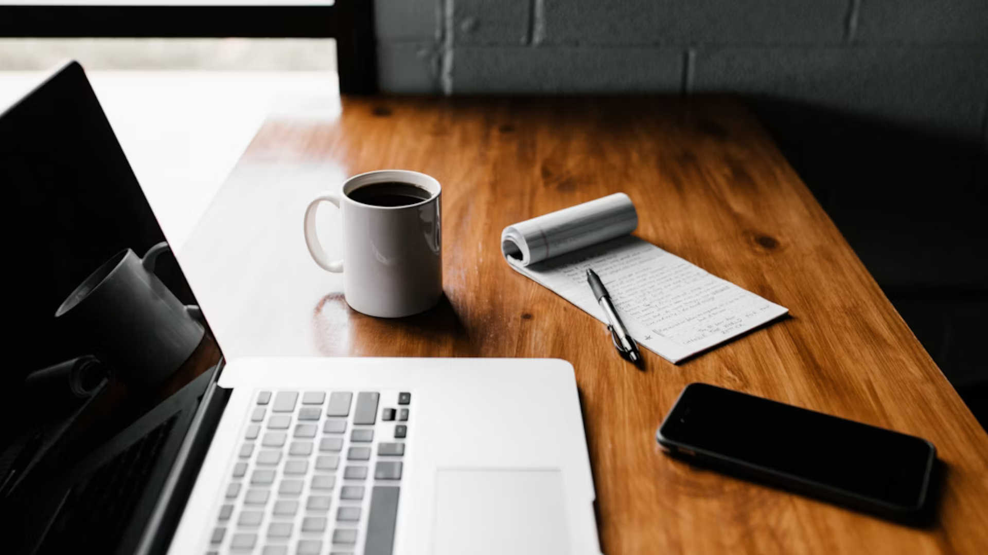 a desk with a computer, phone, coffee and notepad on them